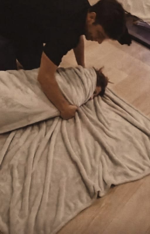 One person kneeling over another who is wrapped up in a blanket on a wooden floor.