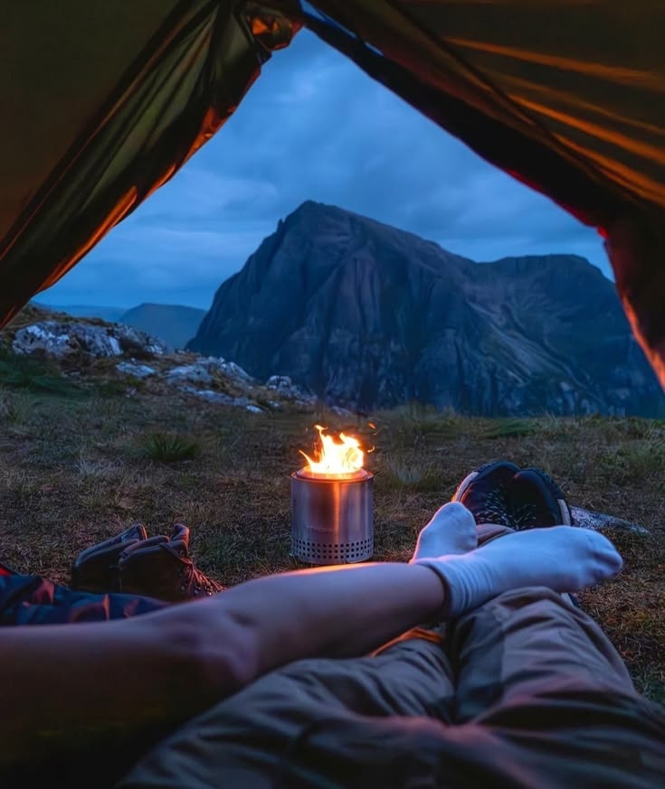 The view from inside a tent at dusk: a campfire, a mountain across the water, somebody's legs in the foreground.