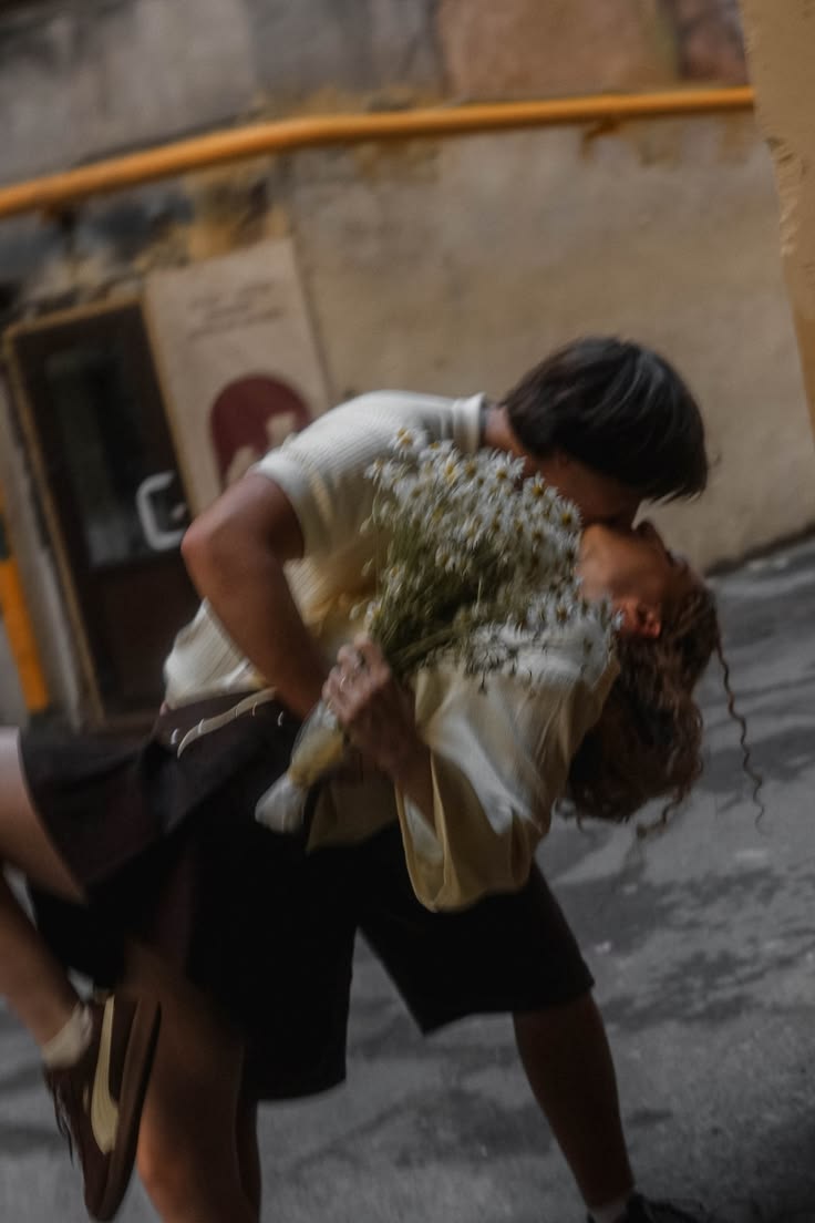 One person carrying the other in their arms on a street, flowers in their hands.
