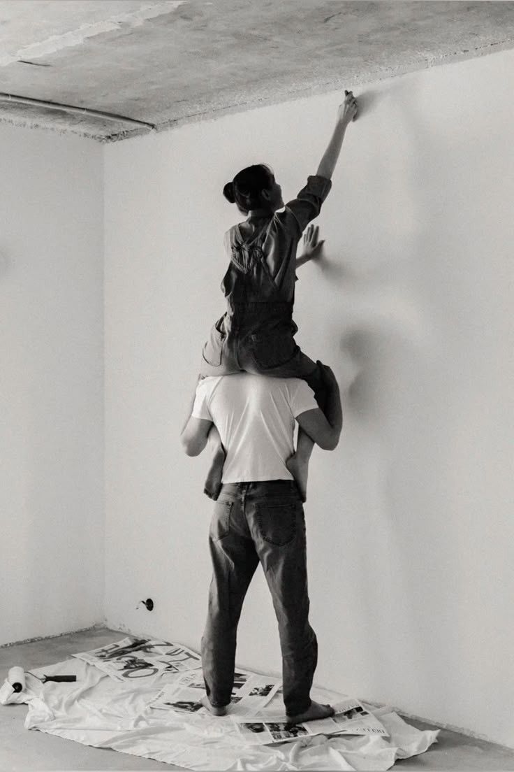 One person sitting on the other's shoulders to paint the wall of an empty room.