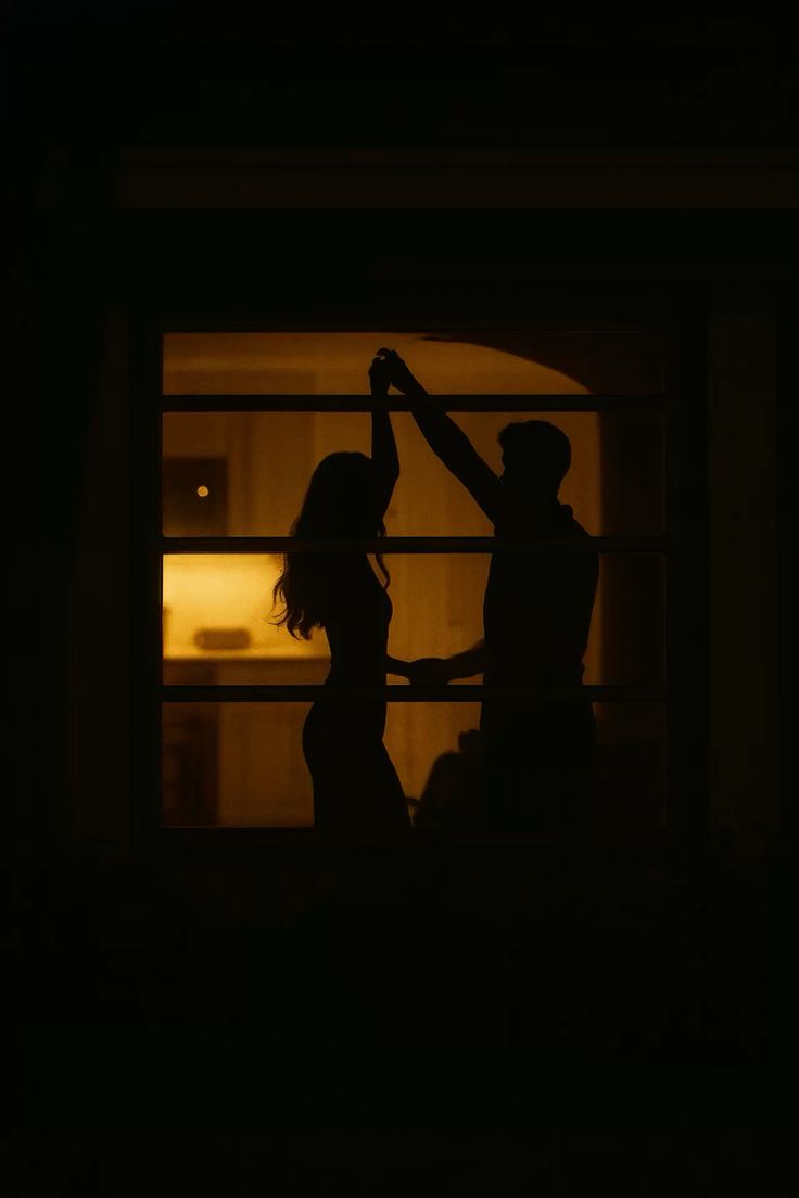 Two people dancing, seen as silhouettes through a lit window at night.