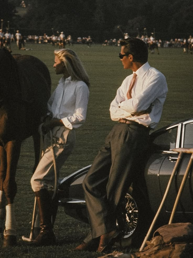 Two people leaning on a rail at a polo field, a horse standing behind them.