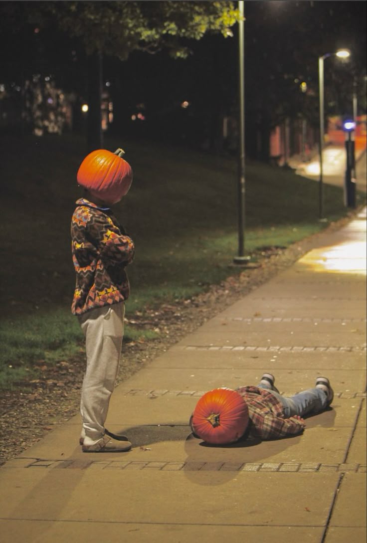 A person standing on a path at night with a carved pumpkin over their head, another lying on the ground beside one.