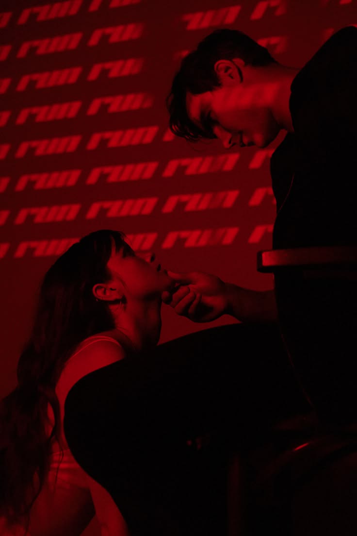 Two people close together in a room lit deep red, lettering across the wall behind them.
